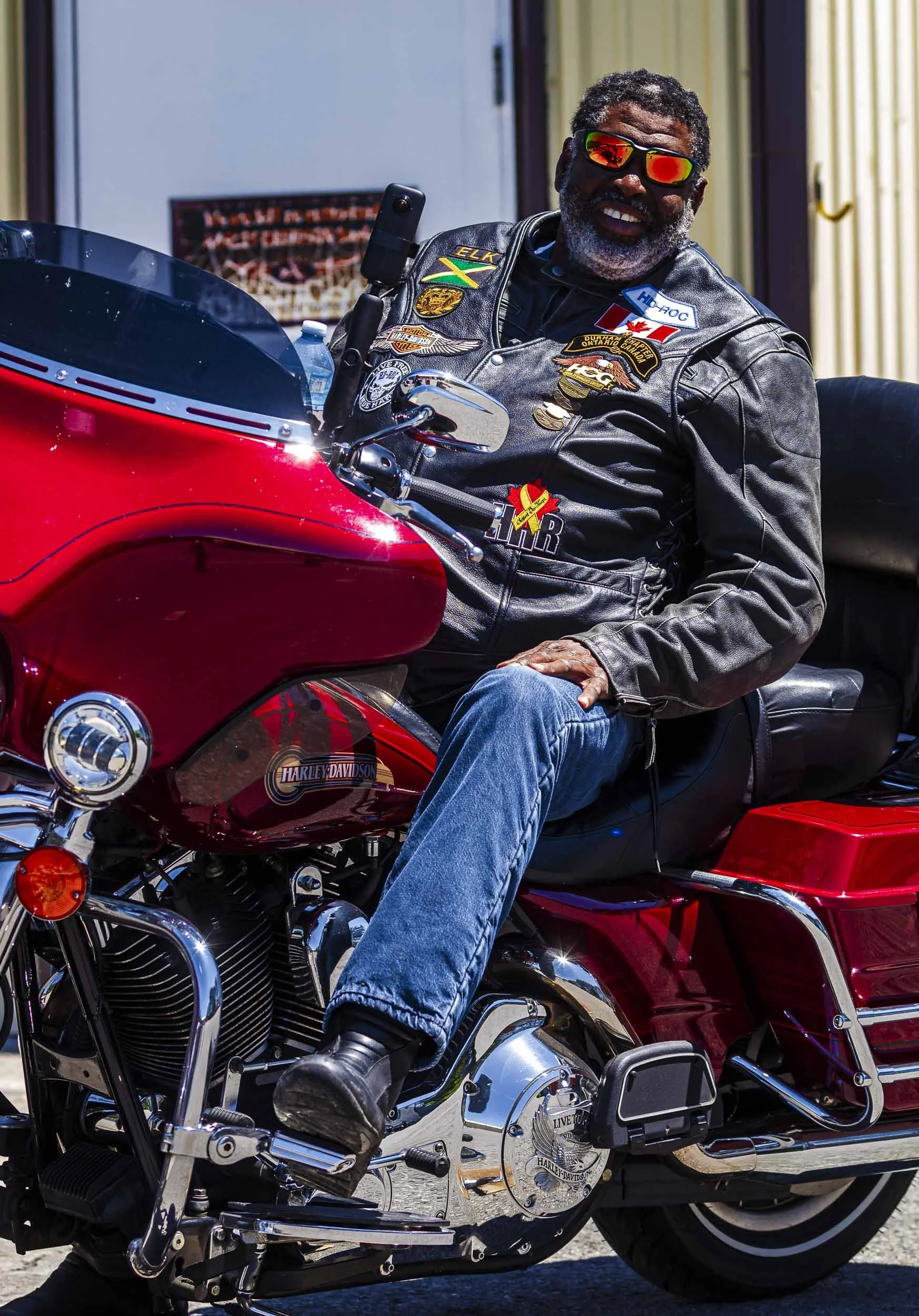 A smiling man wearing sunglasses and a leather vest with Harley Davidson patches, sitting on a red Harley Davidson motorcycle.