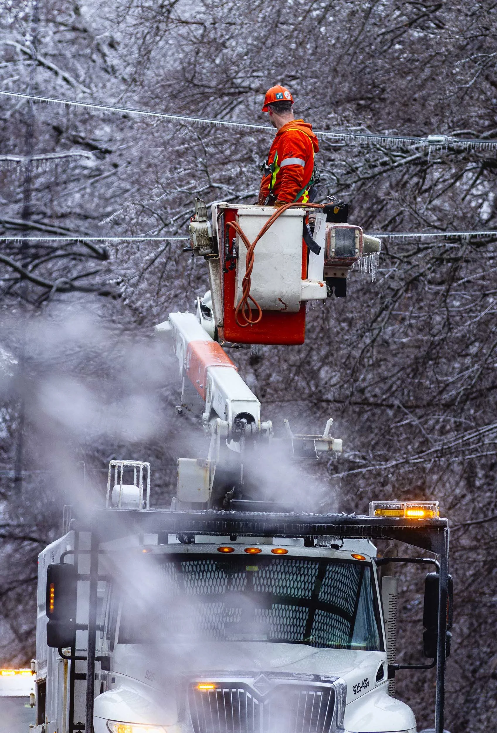 Utility worker in orange safety gear and helmet working on power lines from a cherry picker lift, with a snow-covered background of leafless trees.