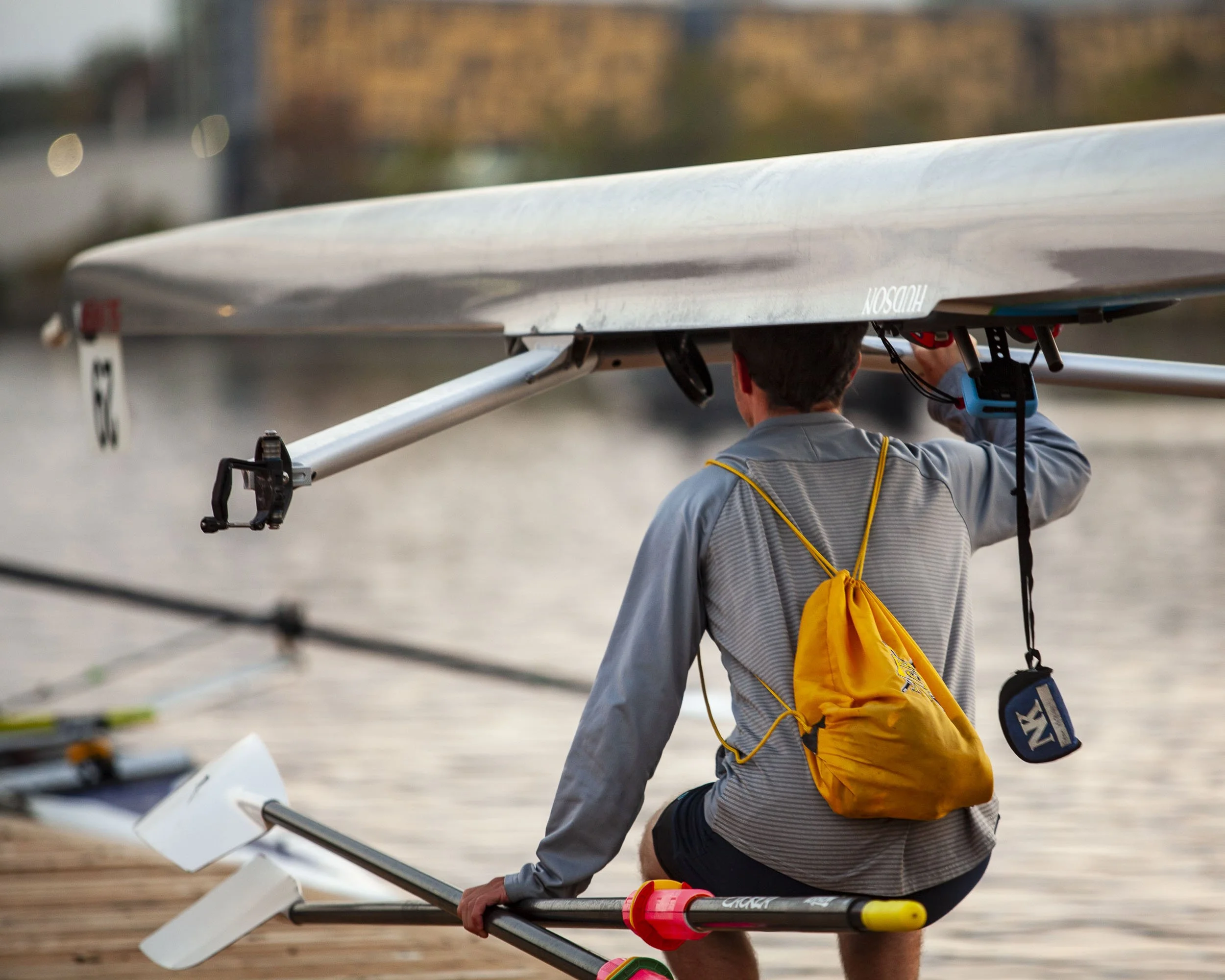 A person in a gray jacket and black shorts sitting on a rowing shell at the water's edge, holding an oar, with a yellow drawstring backpack and small pouch hanging from their shoulders, on a dock near a body of water.