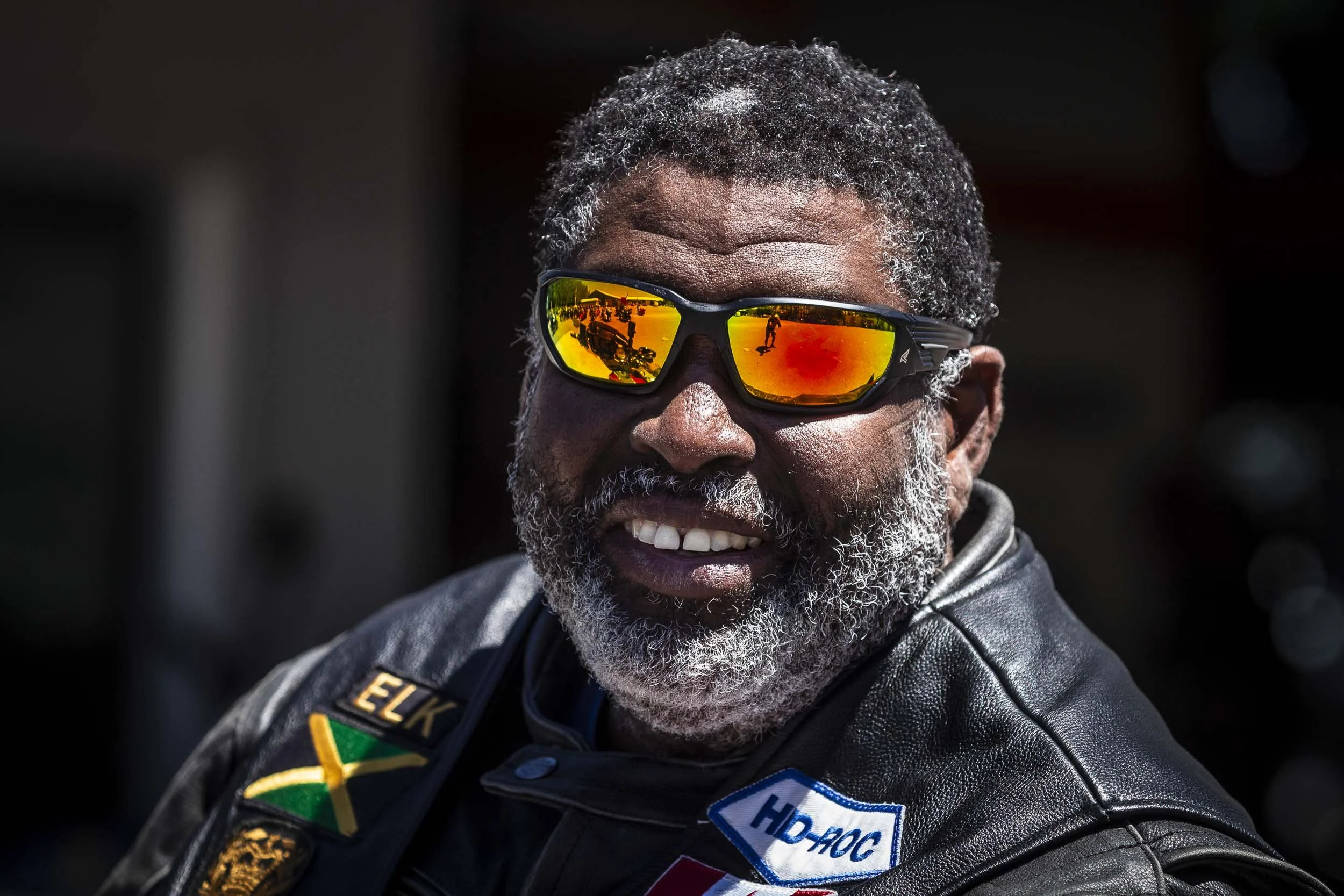 A man with gray hair and beard wearing reflective orange sunglasses and black leather jacket with patches, smiling outdoors.