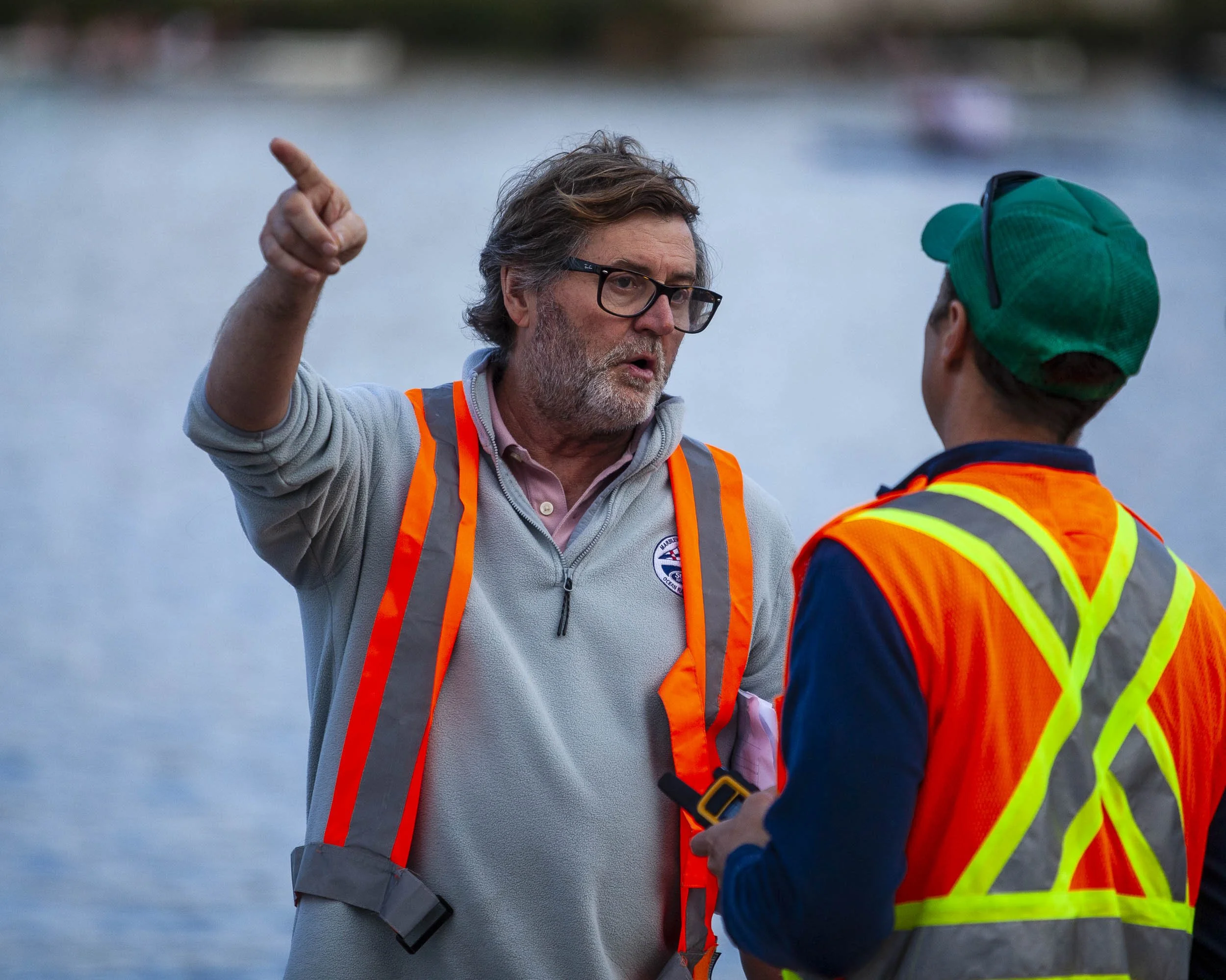 Two men in safety vests having a conversation by a body of water, with one man pointing and gesturing.