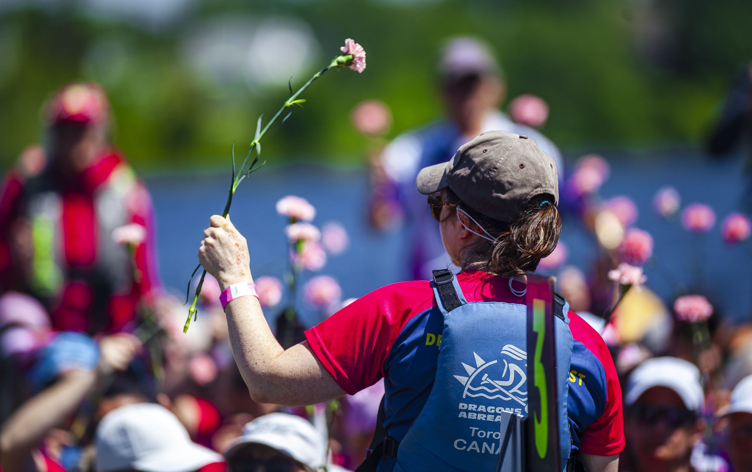A woman in a hat and sunglasses holding a pink flower above a crowd during an outdoor event, with a lake and trees in the background.