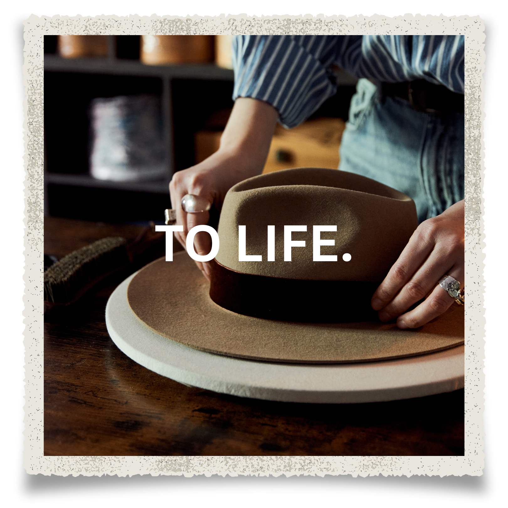 A person wearing a striped shirt is placing a tan fedora hat on a wooden table, with the words 'TO LIFE.' superimposed on the image.