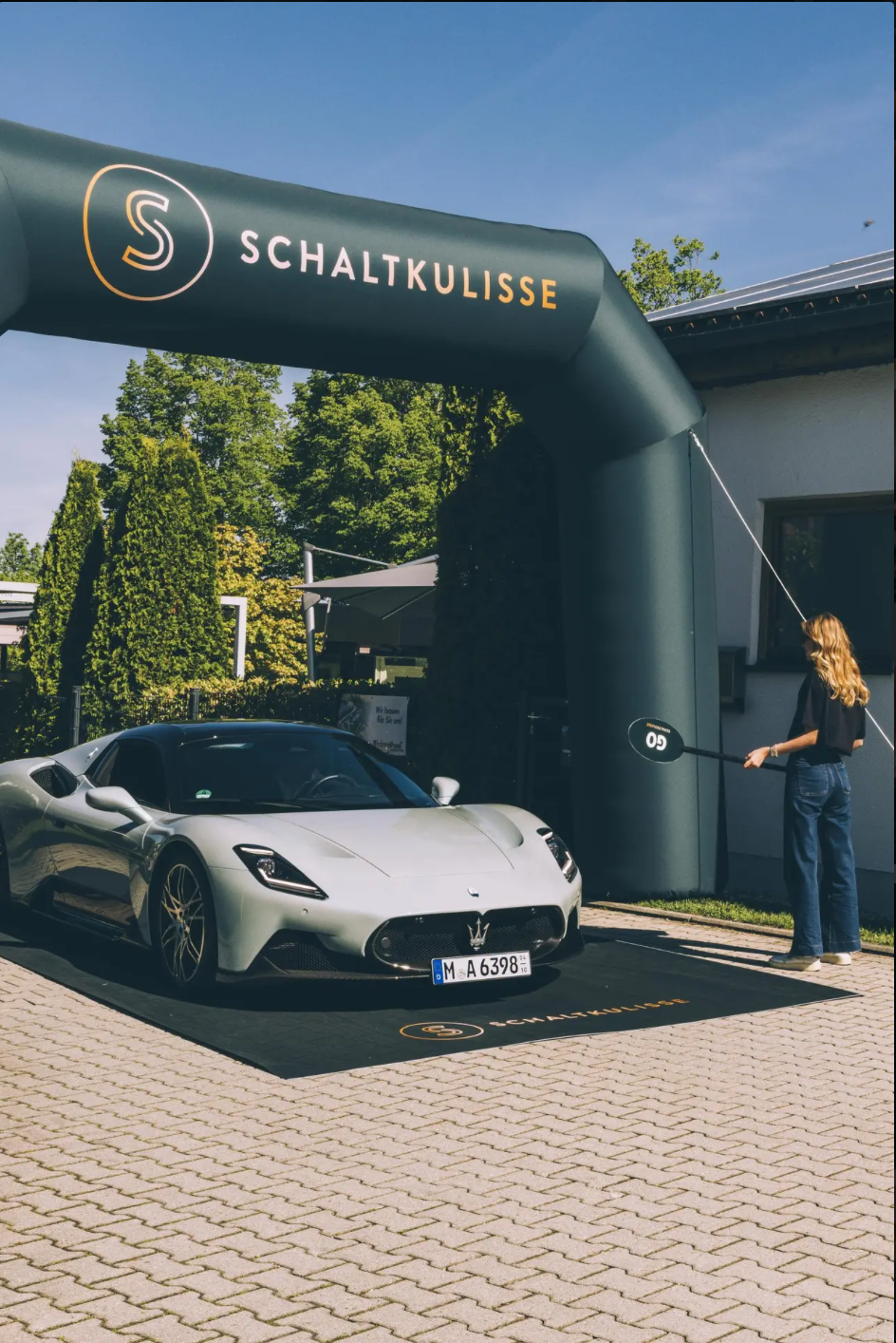 A silver sports car parked under a large green inflatable arch with the logo and the word 'Schaltkulisse'. A woman stands nearby holding a paddle with the number 60 on it, pointing towards the car. The setting appears to be outdoors on a sunny day.