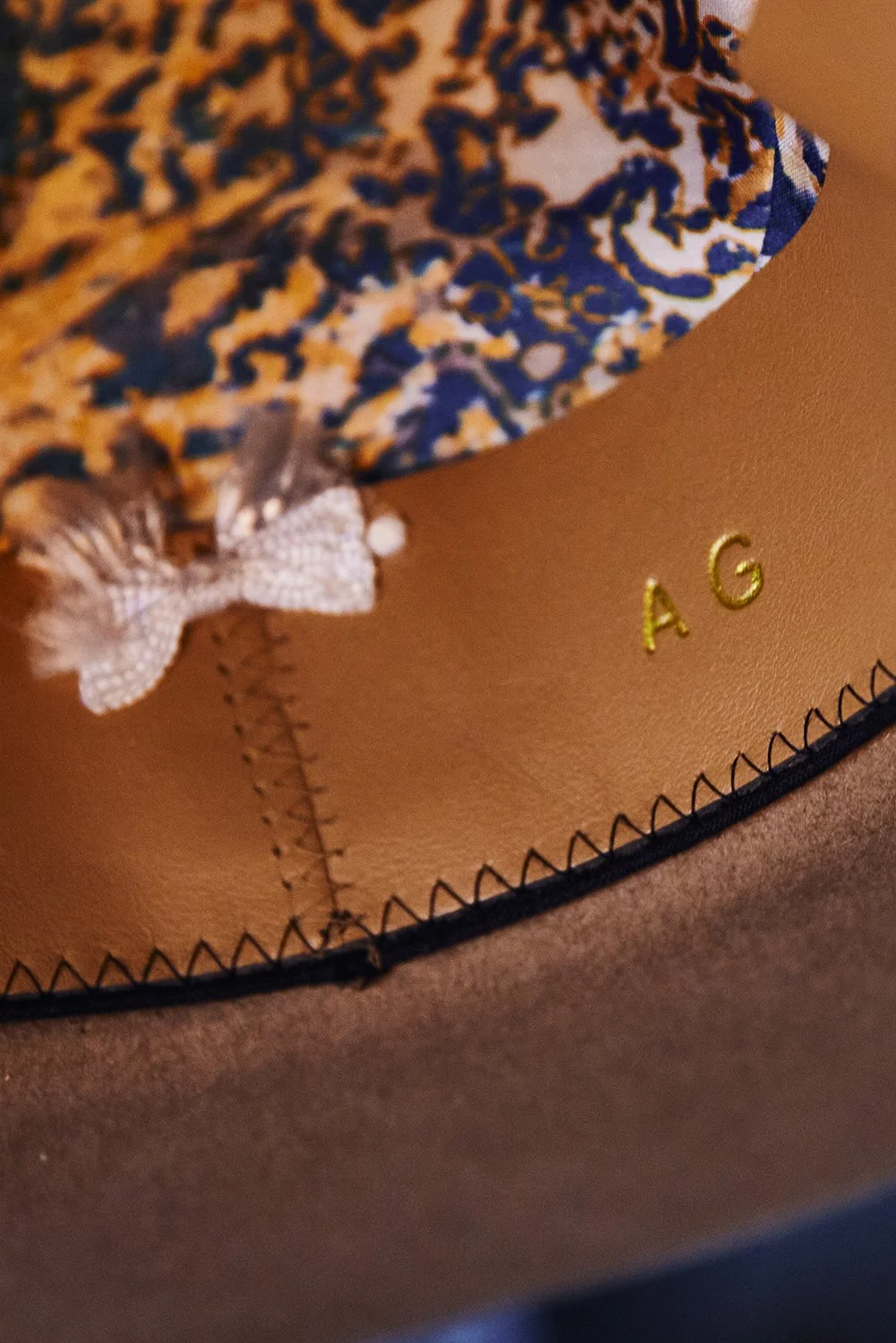 Close-up of a leather item with gold-embossed initials 'A G', showing detailed stitching and a decorative fabric pattern with a butterfly-shaped embellishment.