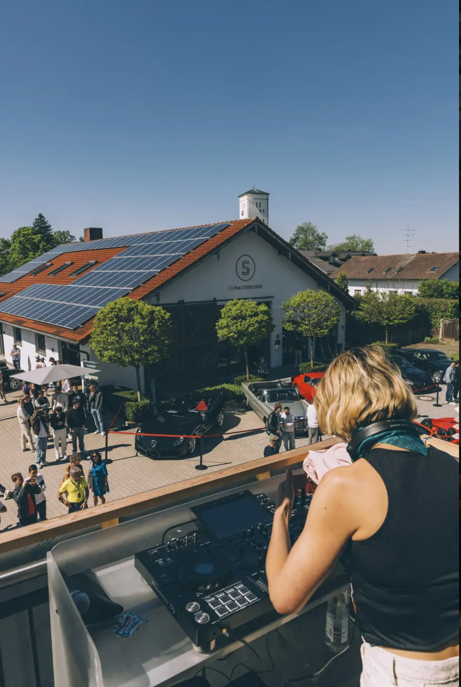A woman DJing at an outdoor event or party on a balcony, overlooking a crowd and a building with solar panels on the roof.