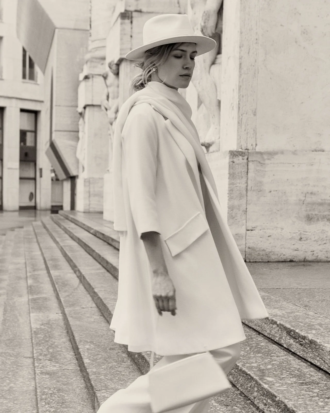 A woman dressed in light-colored, fashionable clothing, including a coat and wide-brimmed hat, walking on a city street with classic architectural details in black and white.