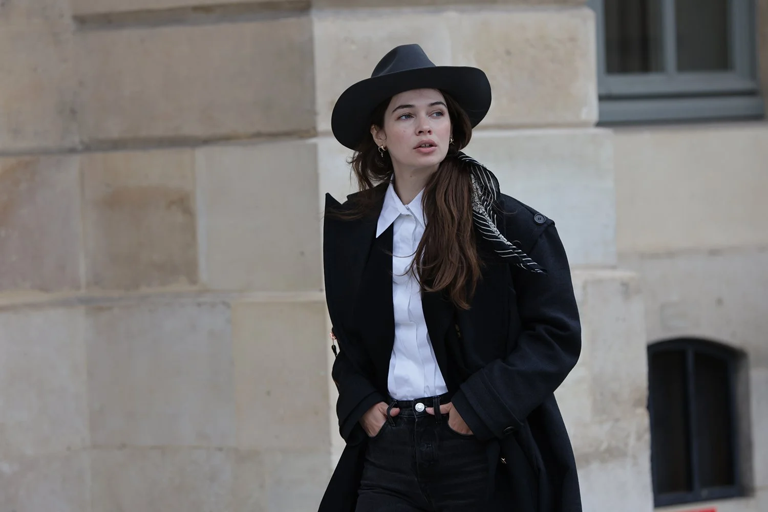 Woman wearing a black hat and coat, standing outdoors in an urban setting.