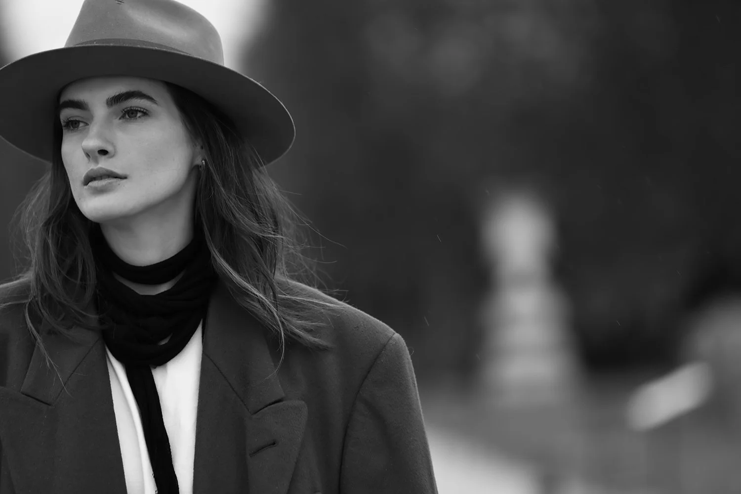Black and white photo of a woman wearing a wide-brim hat  and a blazer, standing outdoors with blurred background.