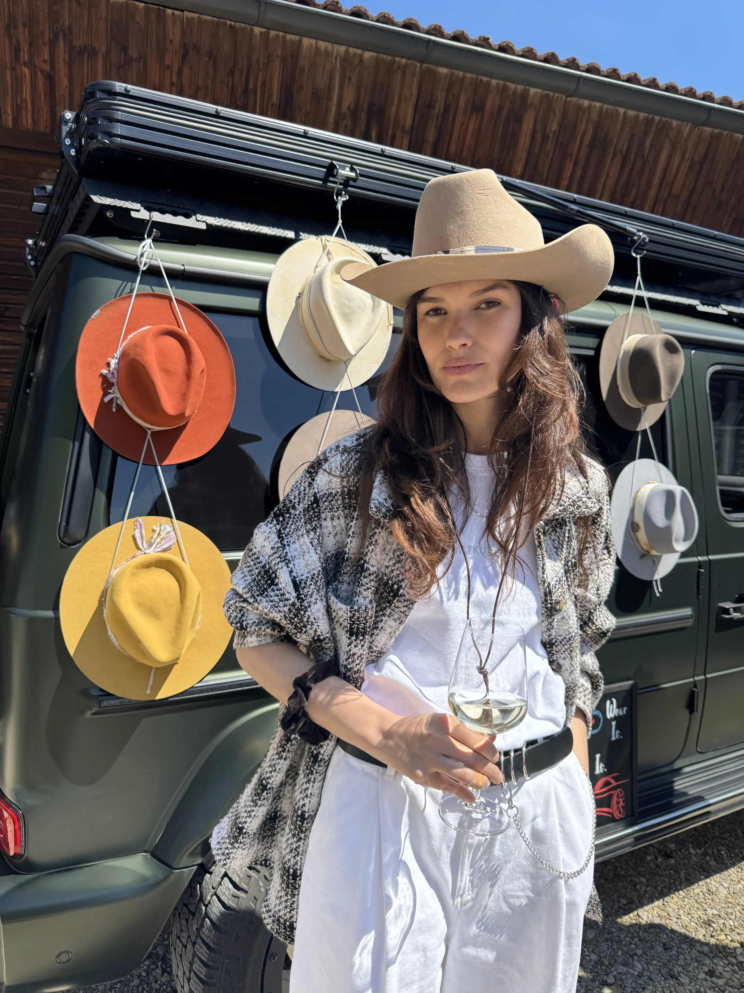 Young woman with long wavy dark hair wearing a beige cowboy hat, plaid jacket, white t-shirt, white pants, and black belt with a chain, holding a glass of white wine. She stands in front of a dark green vehicle with hanging hats, including yellow, orange, beige, and gray, attached to the side, and a wooden barn with a blue sky background.