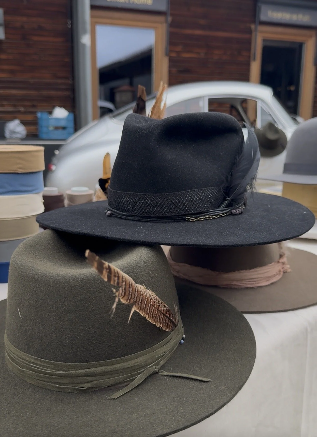 Three wide-brimmed hats with decorative accents, including a feather, displayed outdoors with a vintage white vehicle and a brown wooden building in the background.