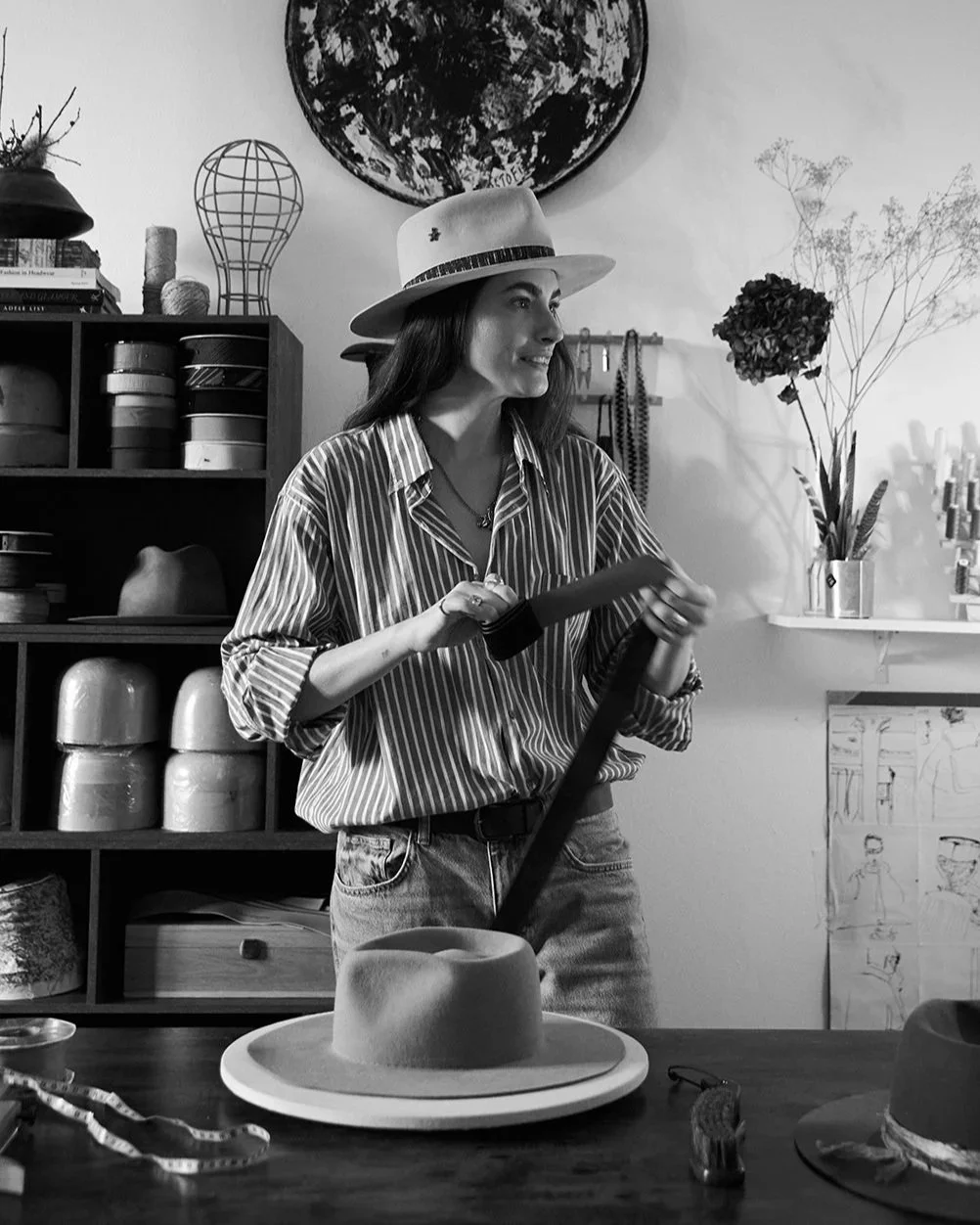 A woman wearing a striped shirt and a hat is standing in a room, holding a measuring tape, with a hat on a table in front of her. The room has shelves with various hats, a large globe wall art, and plants.