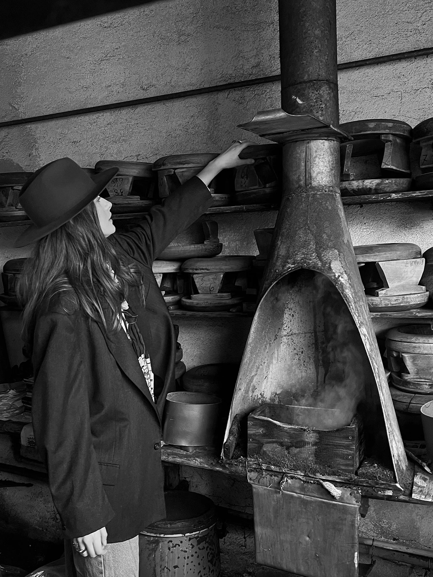 A woman wearing a wide-brimmed hat and blazer standing in front of a vintage wood stove, reaching up to a shelf with stacked cast iron cookware.