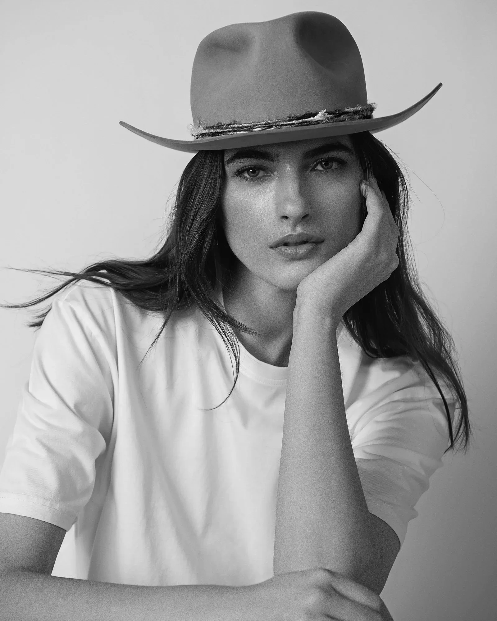 A woman wearing a wide-brimmed hat and a white t-shirt, resting her face on her hand, looking directly at the camera in black and white.