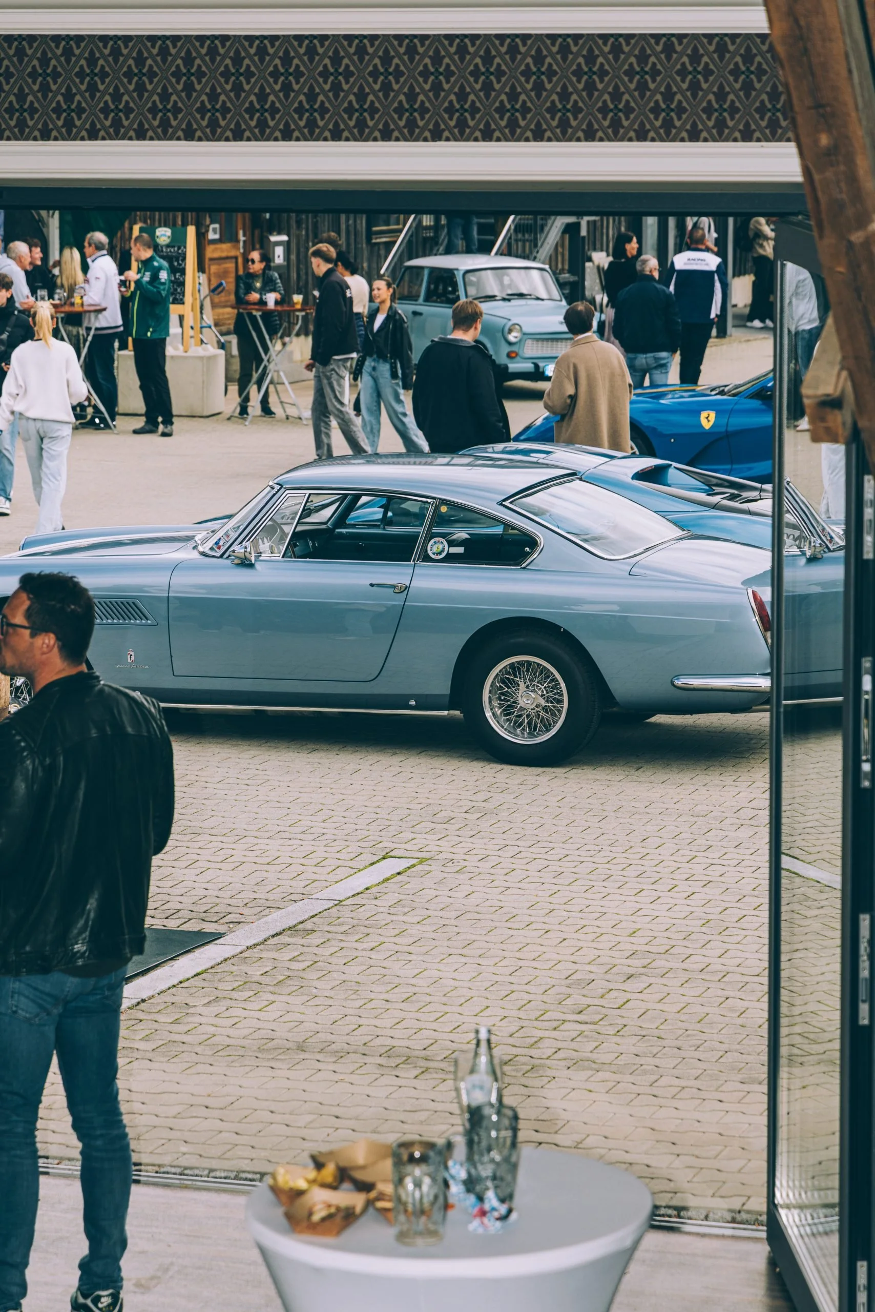 A vintage silver-blue classic car displayed at a car show with several people walking and socializing around it.