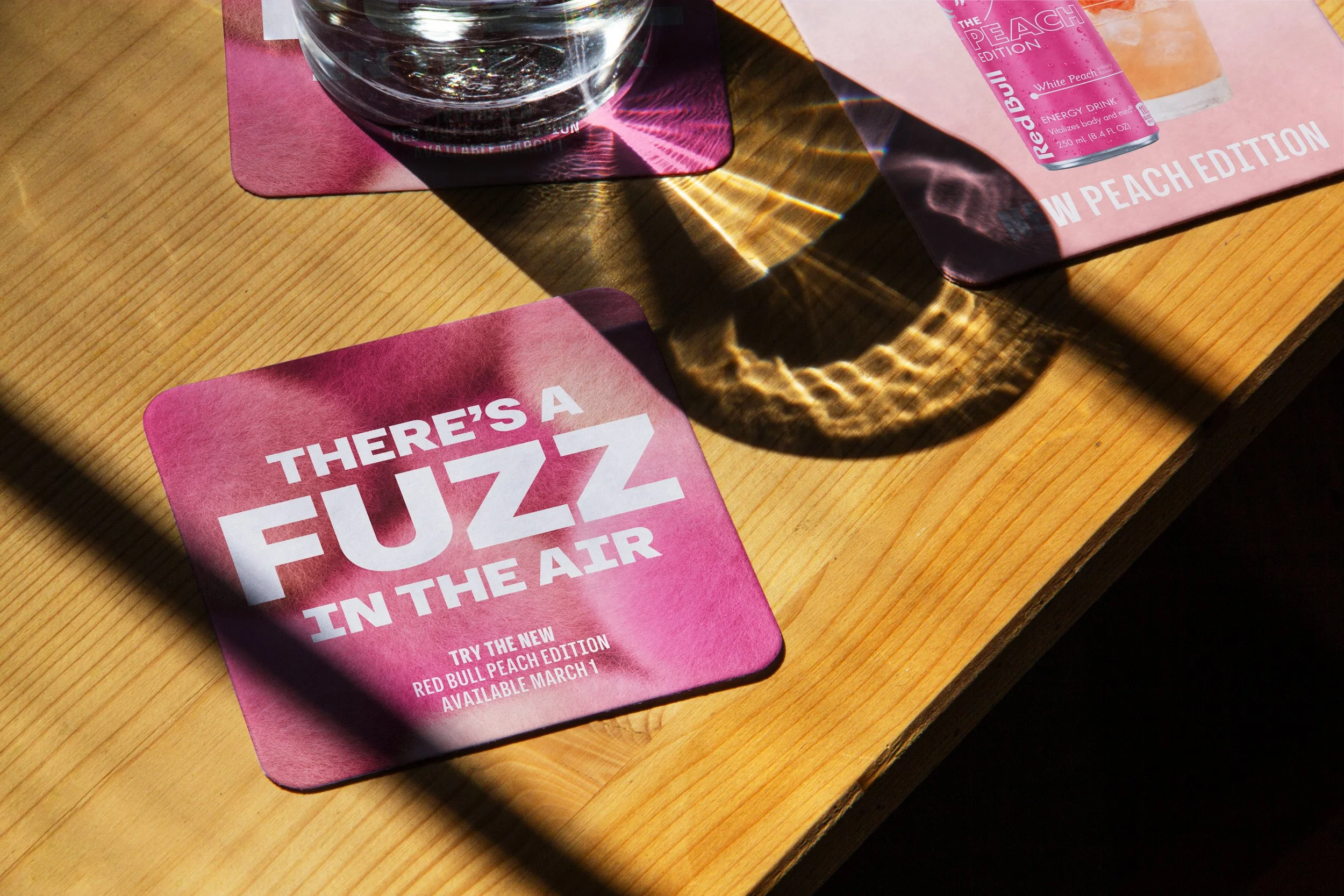 Red Bull Peach Edition coasters on a sunlit wooden bar, featuring the headline ‘There’s a Fuzz in the Air’ against a fuzzy pink background.