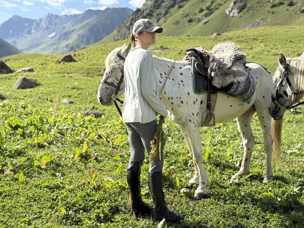 A woman standing beside a saddled spotted Chaar horse on a mountain trail