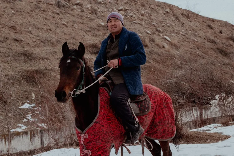 A Kyrgyz rider in a blue coat on a horse with a red blanket