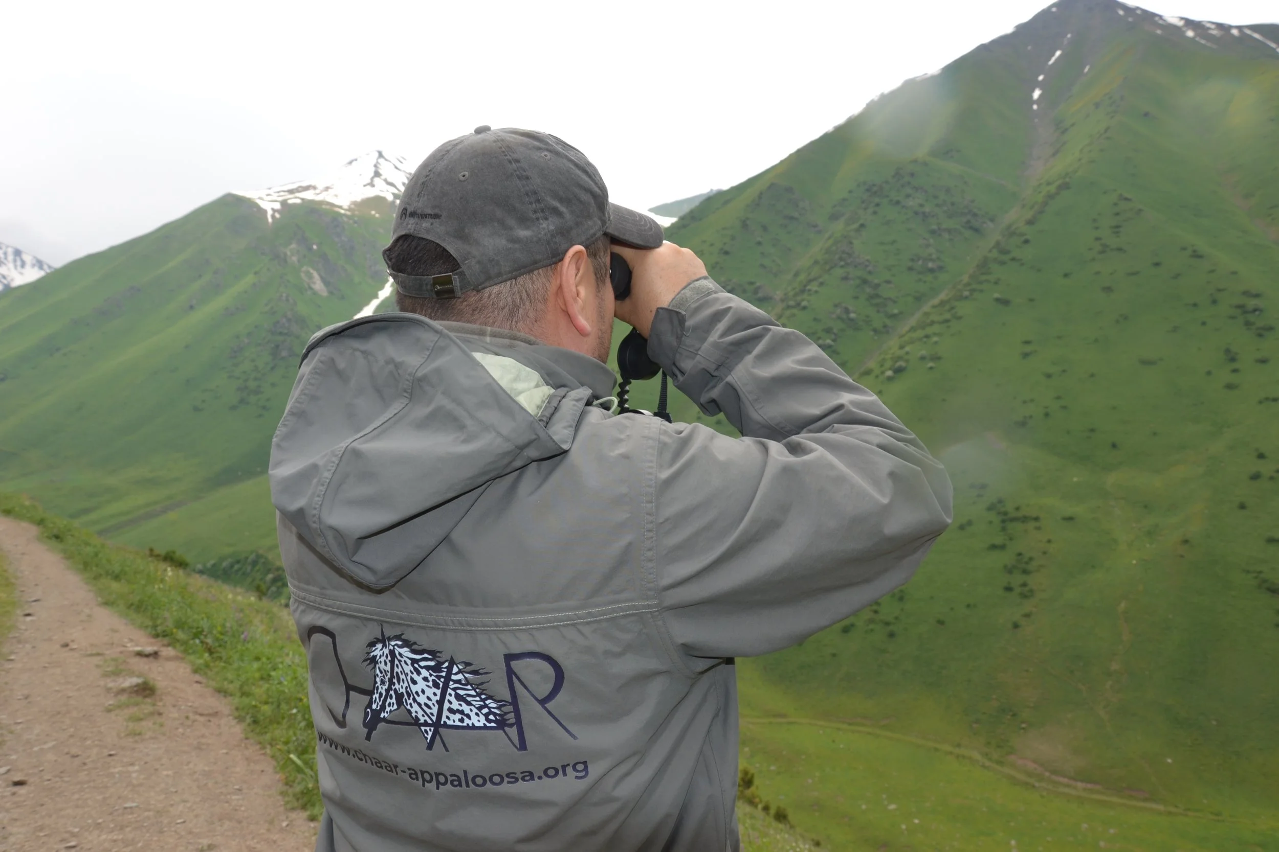 A man in a Chaar jacket looking through binoculars at green mountains