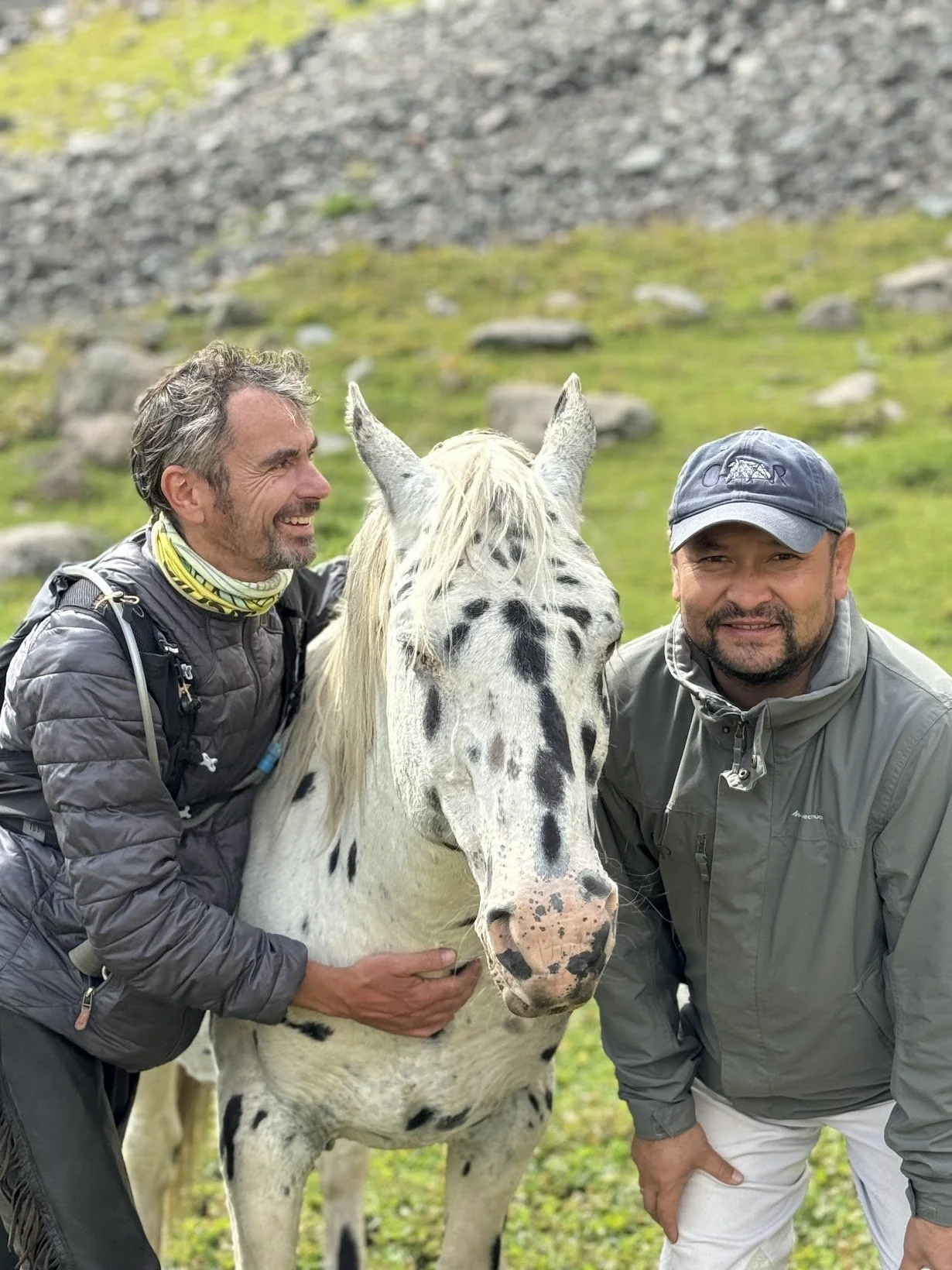 Deux hommes sourient ensemble avec un cheval Chaar tacheté dans un pâturage verdoyant
