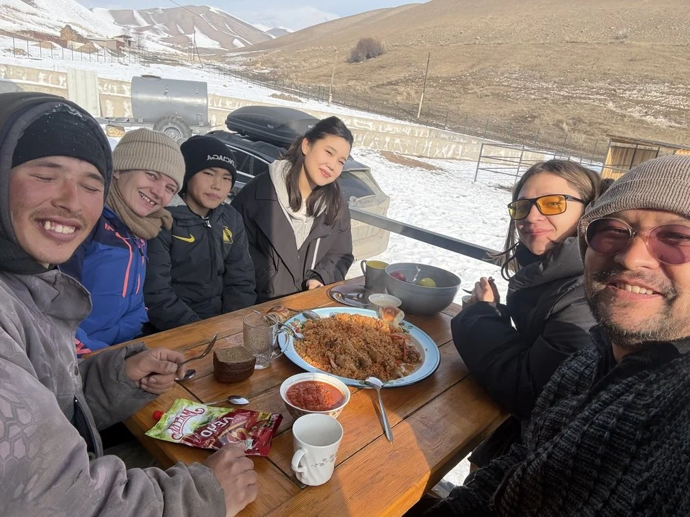 Un groupe partage un repas à une table en terrasse, avec des collines enneigées en arrière-plan