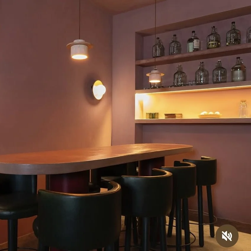 Interior of a bar or cafe with a wooden counter, black bar stools, wall-mounted pendant lights, and glass bottles on shelves, illuminated by warm lighting.