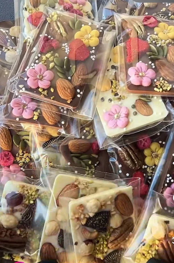 Assorted decorated chocolates wrapped in clear plastic, topped with colorful flower, raspberry, nuts, and other edible decorations.