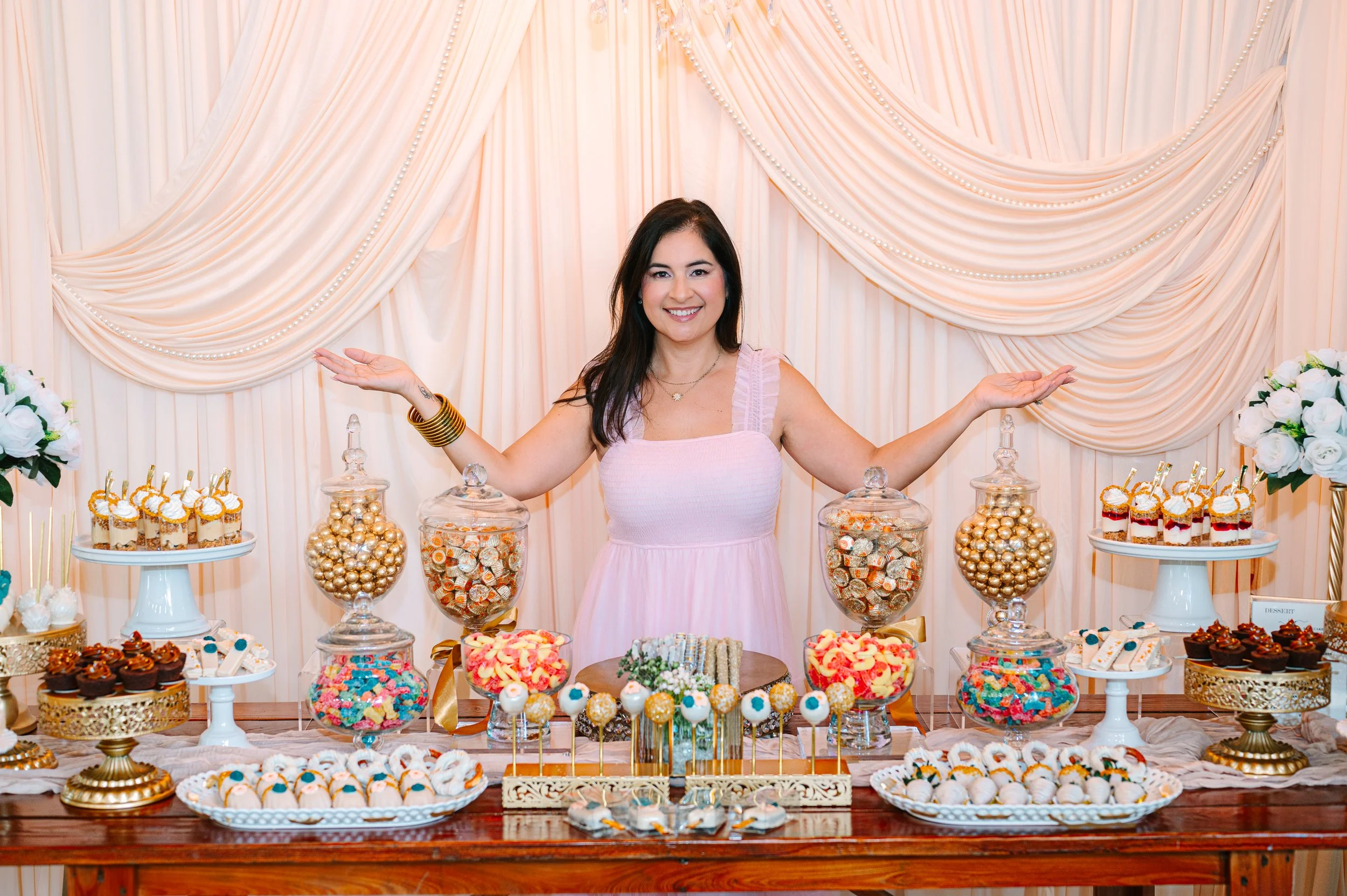 Woman smiling at a dessert table decorated with pink and gold themed treats, including cupcakes, candies, and cookies, against a pink draped backdrop with flowers.