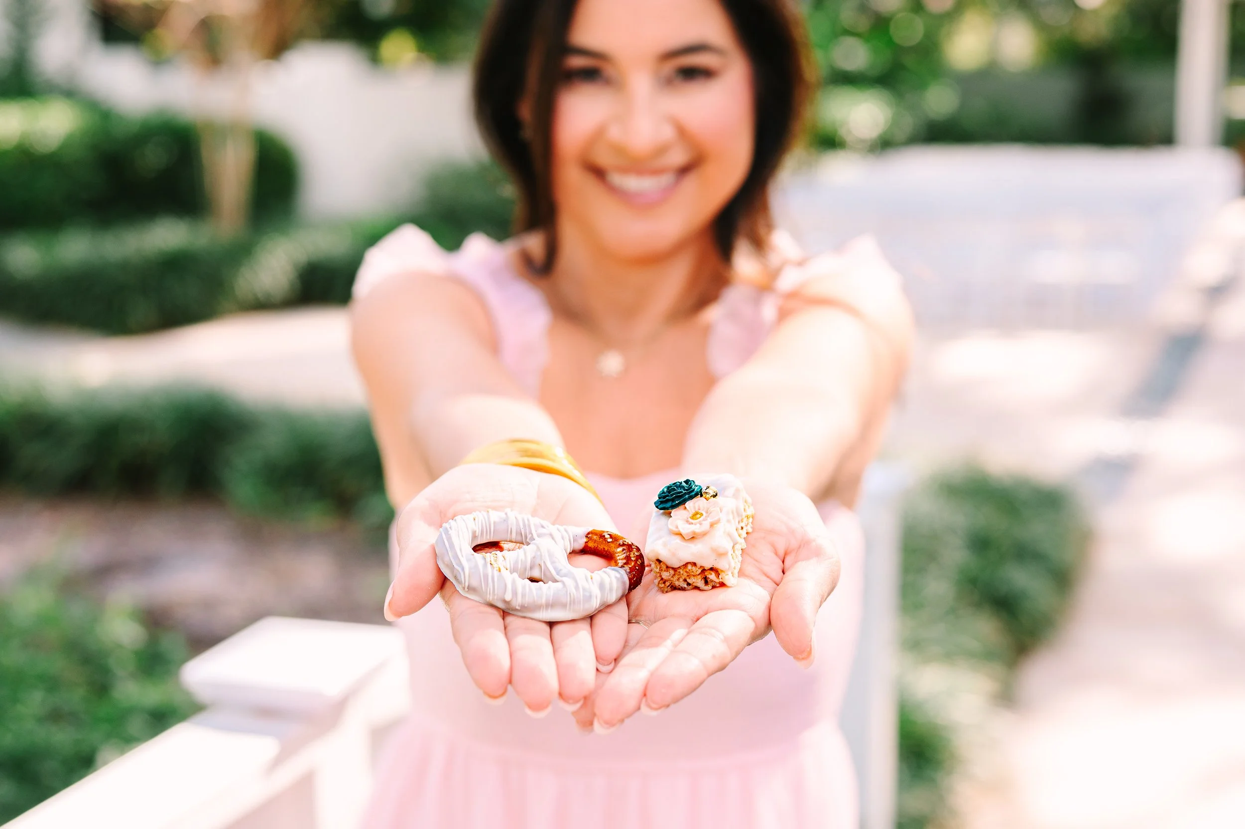 A woman in a pink dress holds out her hands showing a pretzel and a pastry with decorative flowers outside on a sunny day.