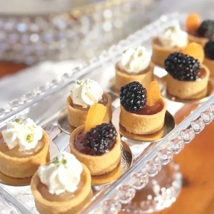 Miniature fruit tarts topped with blackberries, slices of peach, and whipped cream, displayed on a glass tray.
