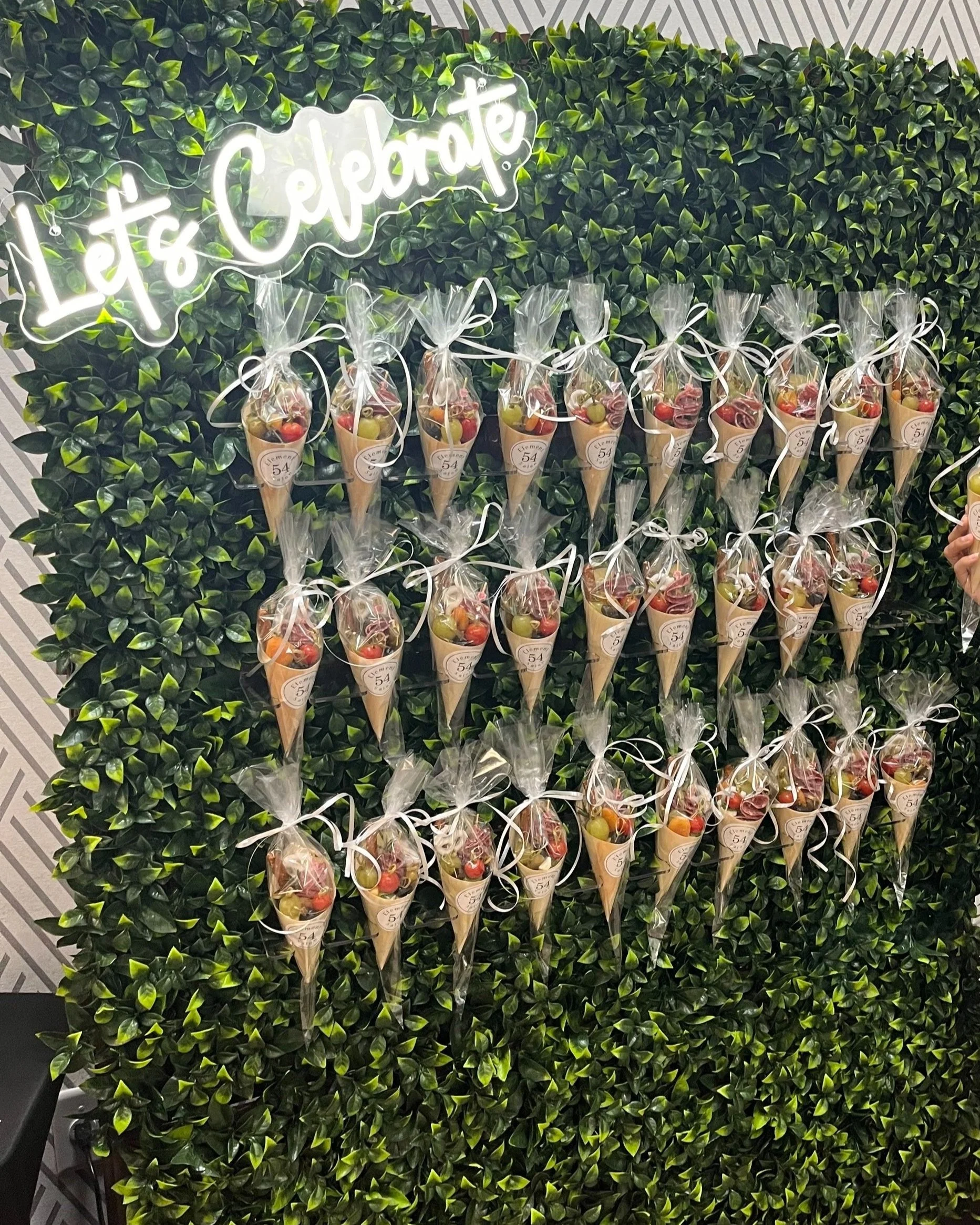 Green leafy wall with a neon sign that says 'Let's Celebrate' and numerous small cones filled with fruit, wrapped in clear plastic with white ribbon.