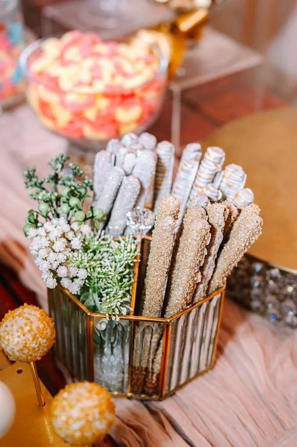 A metallic gold striped container filled with various decorated cookies and sweets, with a small green plant and white flowers, set on a pink tablecloth. In the background, there is a bowl of pink and white candies.