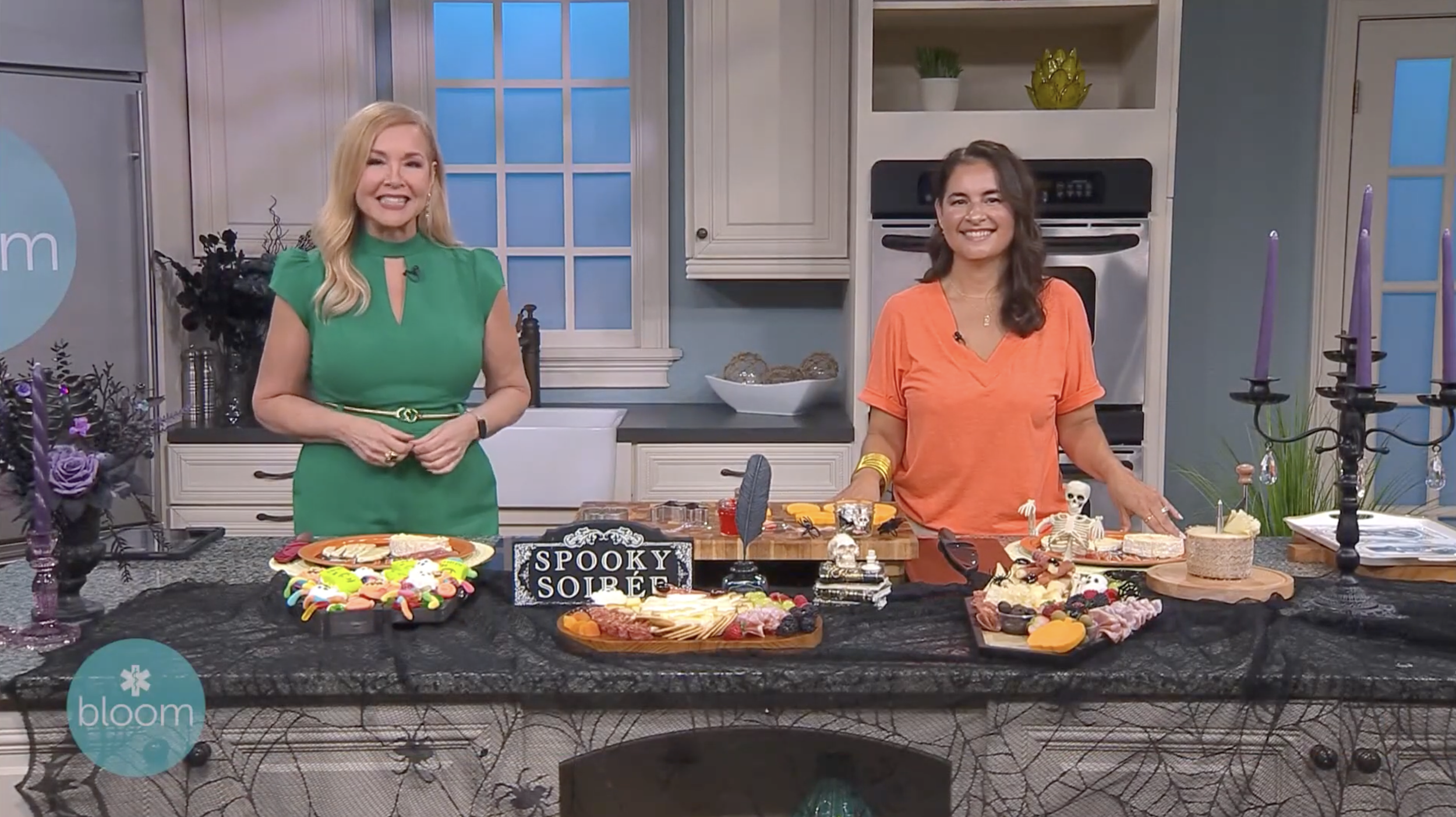 Two women presenting Halloween-themed snacks on a kitchen counter decorated with a black spiderweb cloth, purple candlesticks, and skeleton decorations, with a blue window and shelves in the background.