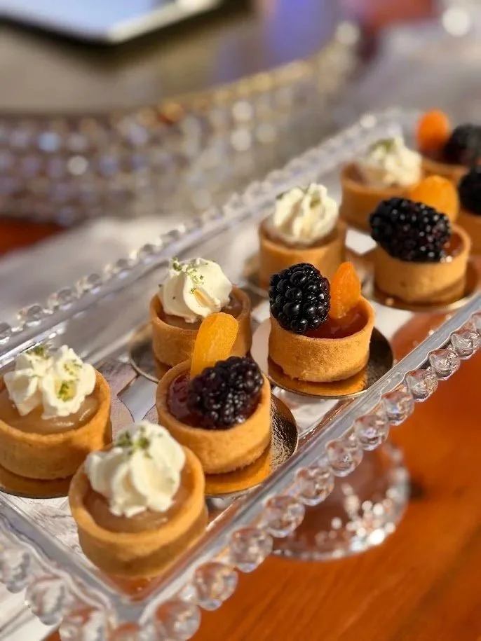 Mini fruit tarts topped with blackberries, dollops of whipped cream, and small orange fruit slices on a glass platter.