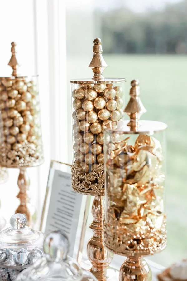 Three tall glass containers filled with gold and bronze decorative balls and wrapped candies, each topped with a decorative gold finial, on a table near a window.