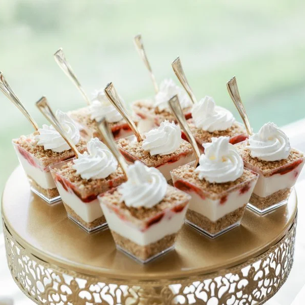 Eight individual layered dessert cups with whipped cream, graham cracker crumbs, and strawberry or raspberry topping, garnished with small silver forks, arranged on a decorative gold cake stand.