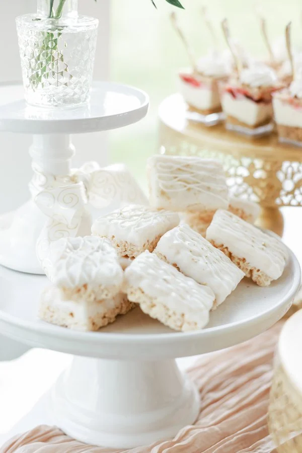 Assorted white frosted rice crispy treats on a white cake stand at a dessert table.