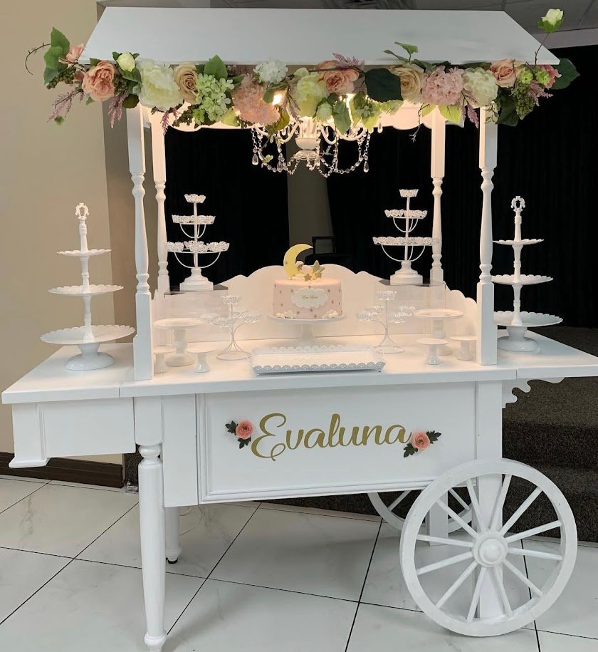 A white dessert cart decorated with pink and white flowers, featuring a cake with moon and star toppers and the name 'Evaluna' on the side, set for a celebration or party.