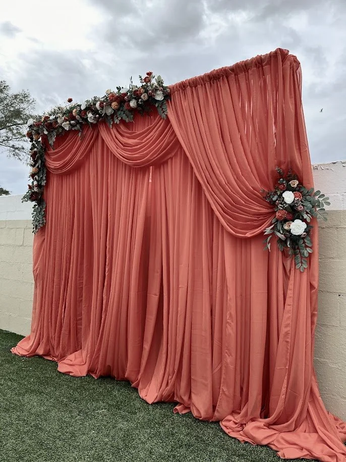 Coral-colored draped fabric backdrop decorated with floral arrangements for an outdoor event.