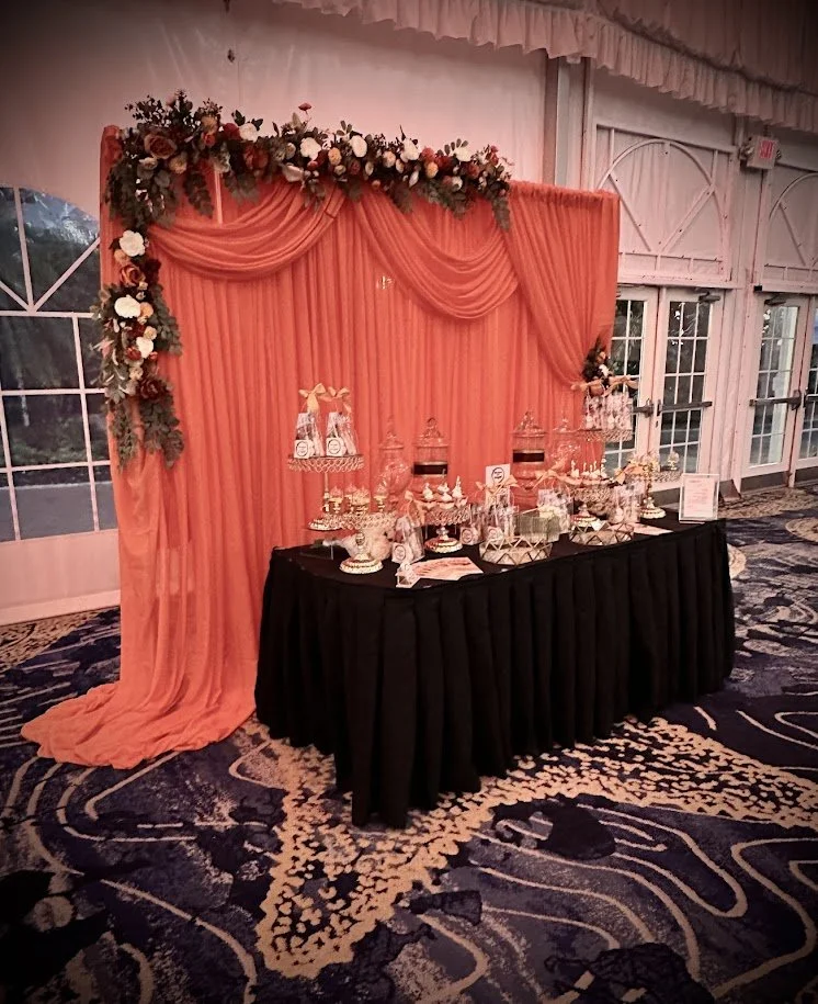 Decorative dessert table with pink drapery and floral arch at an indoor event.