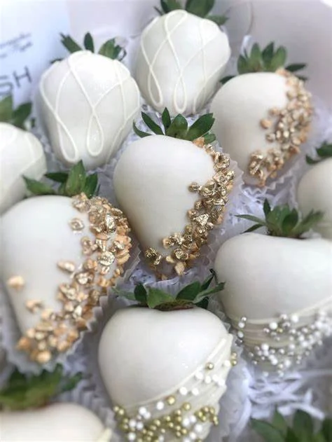 White chocolate-covered strawberries decorated with pearls and gold sprinkles on green leaves.