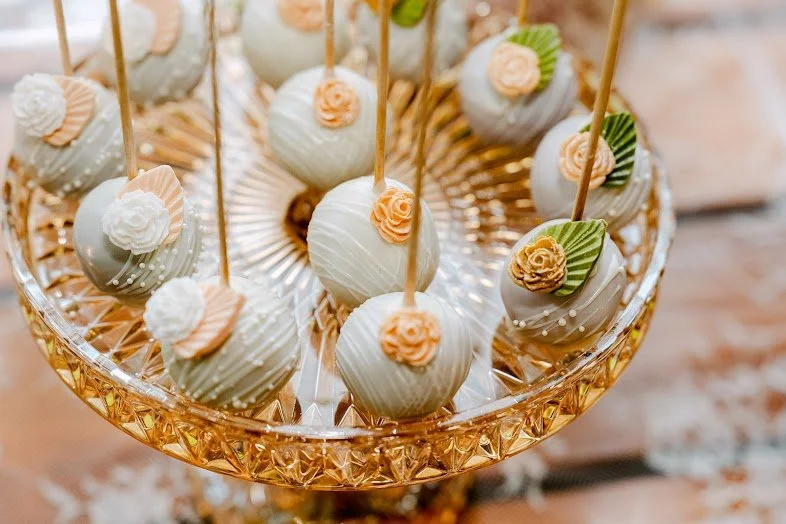 Cake pops decorated with white icing, small peach-colored roses, green leaves, arranged in a glass cake stand.