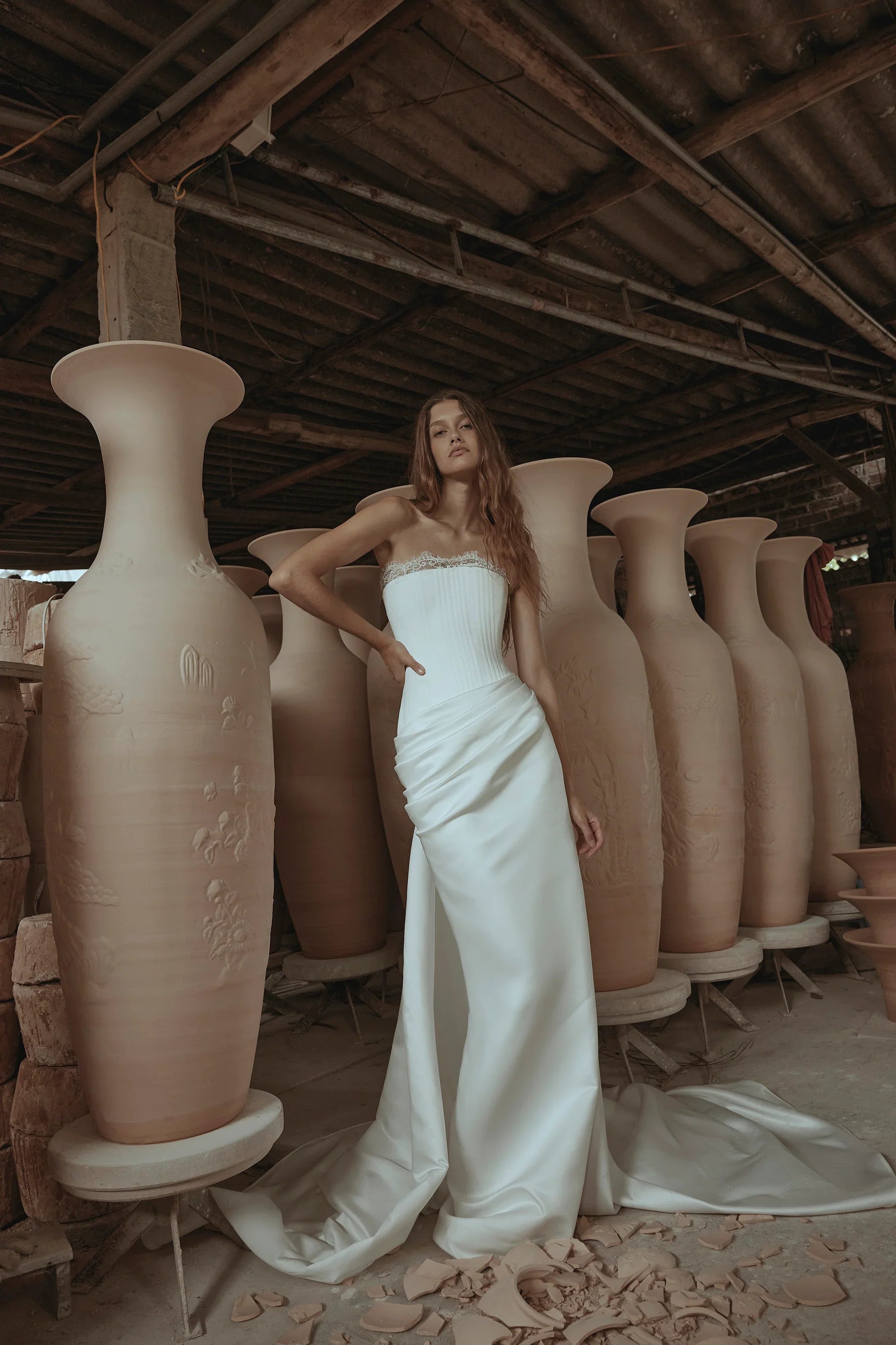 A woman in a white wedding dress standing among large ceramic vases in a pottery workshop.