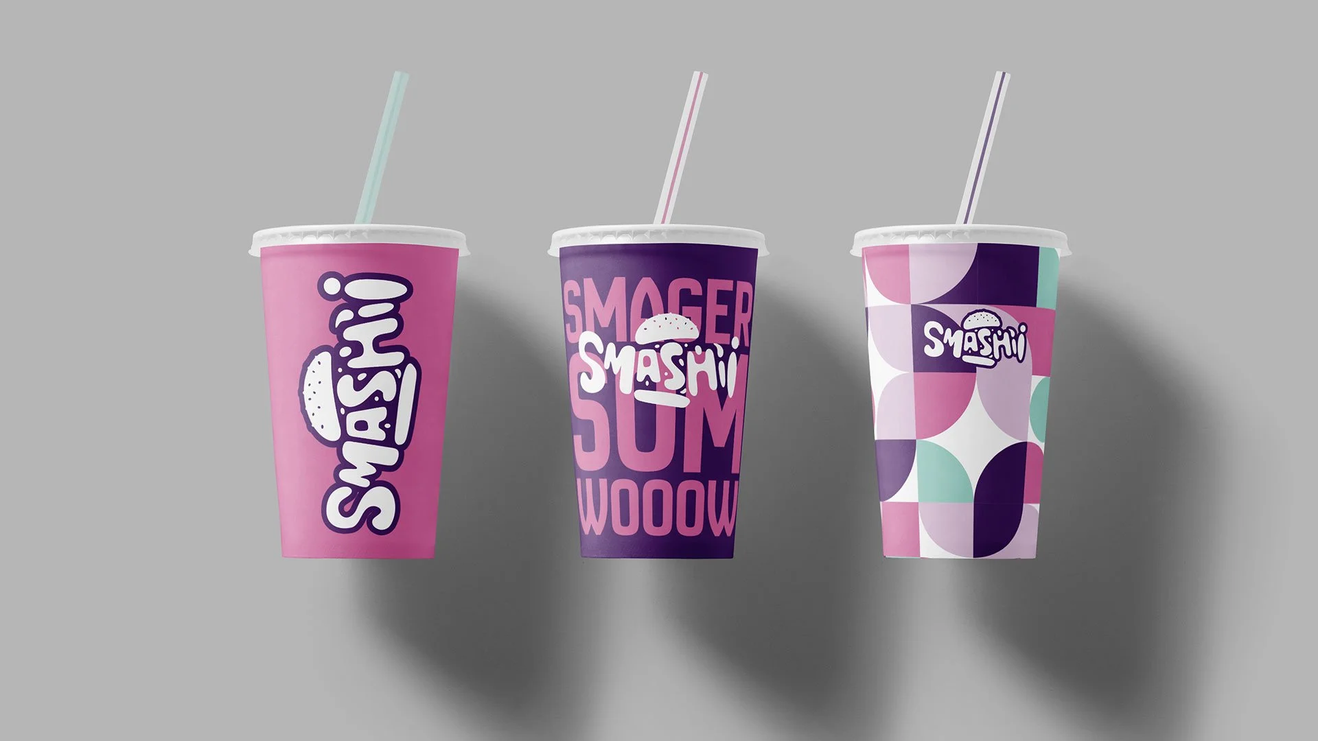 Tre farverige kopper med strå. Den første er pink med logoet 'SMASH!'. Den anden er lilla med teksten 'SMAGER SMASHI SUM WOOW'. Den tredje er mønstret med cirkler i lilla, pink, hvid og grøn med logoet 'SMASHI'.