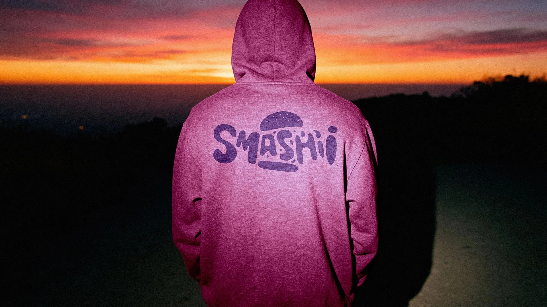 Person i pink hættetrøje med 'SMASHI' logo, set bagfra, står på en bakke under en solnedgang.