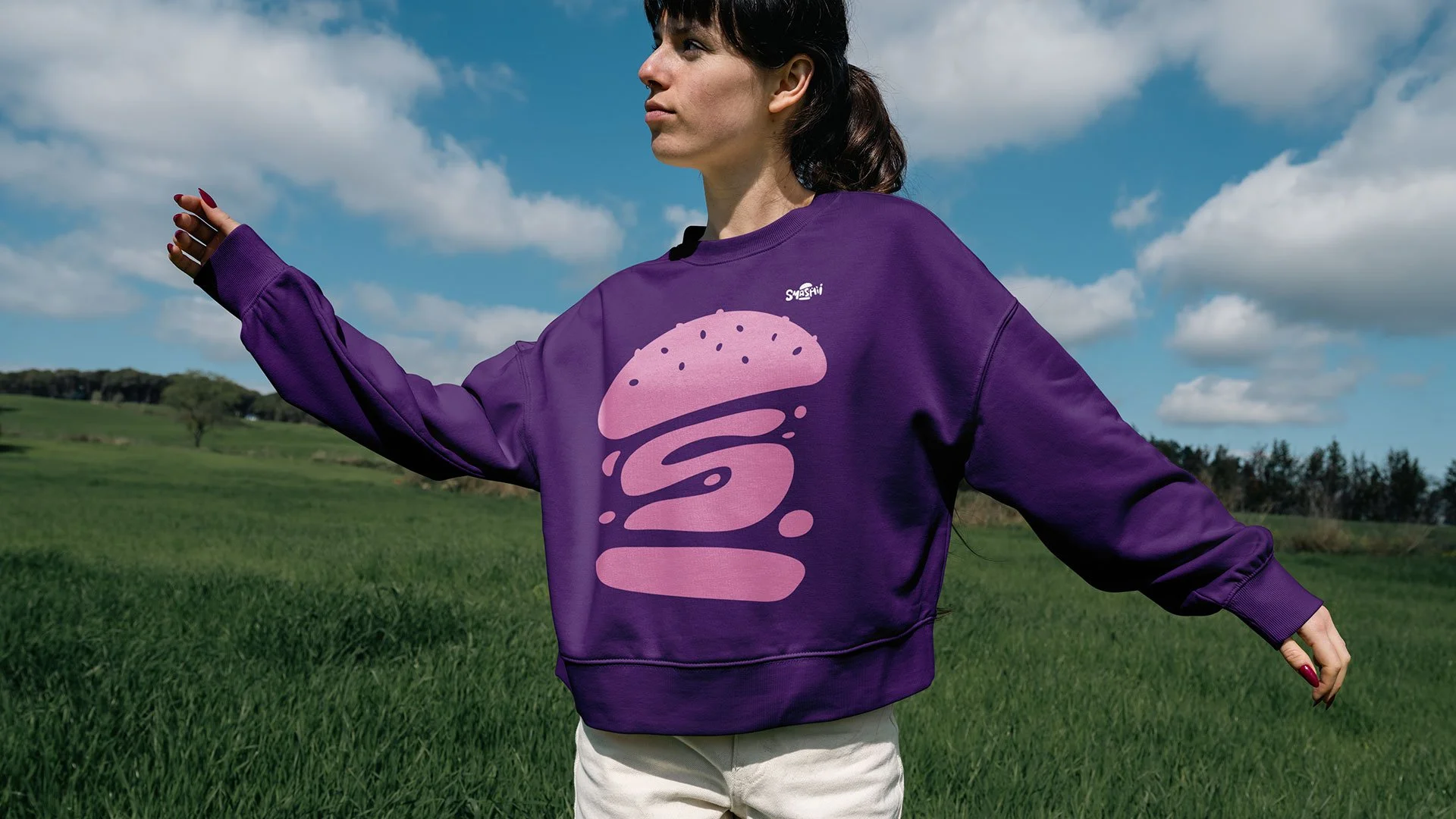 Kvindelig person i en lilla sweatshirt med en pink grafisk design står i en grøn mark under en blå himmel med skyer.
