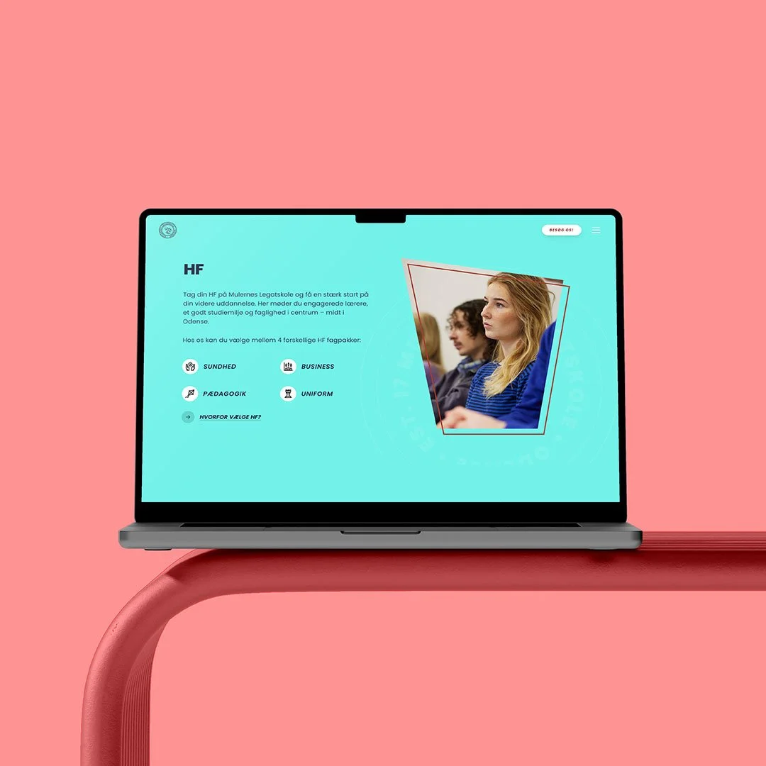 En open laptop på en pink bord, der viser en hjemmeside med tekst og et billede af nogle unge mennesker i et klasseværelse.