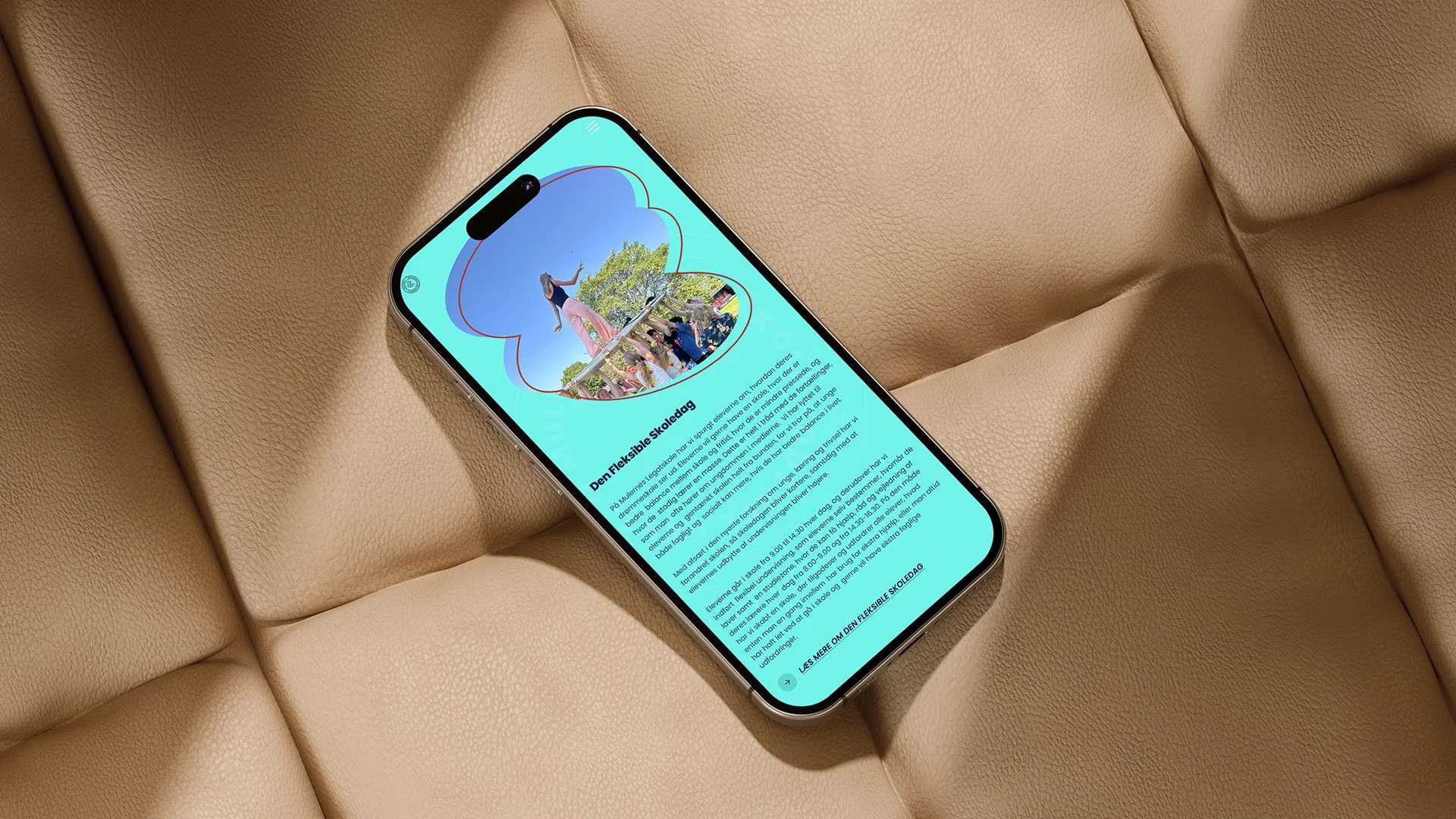 En smartphone ligger på en beige lædersofa og viser en nyhed eller artikel om en fleksibel skoledag med et billede af børn og en voksen, der leger udendørs under en blå himmel.
