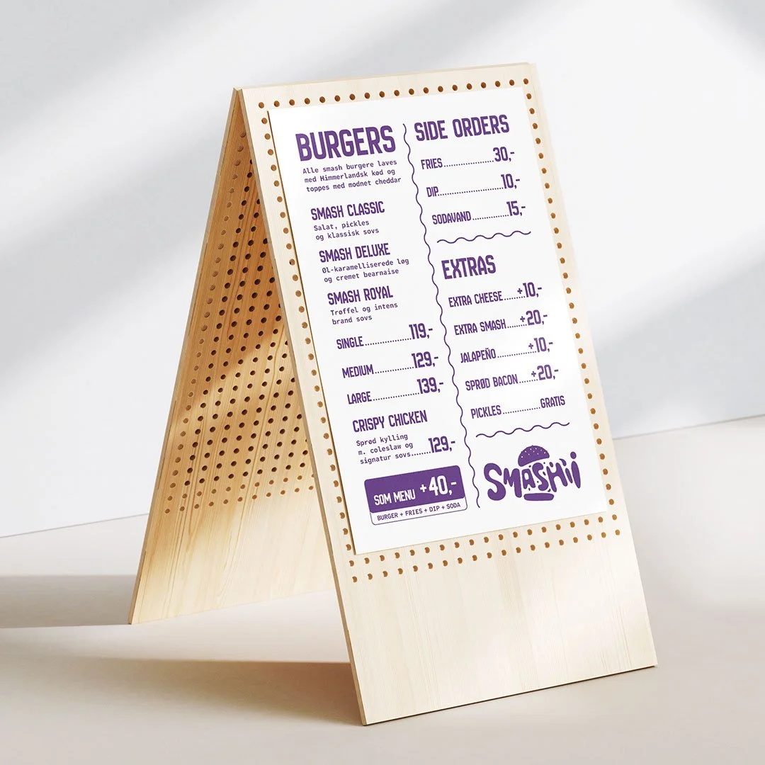 Bord med menukort for burgerrestaurant, der tilbyder forskellige burgervarianter, sideorders, ekstra toppings og tilbehør. Alt er skrevet på dansk.