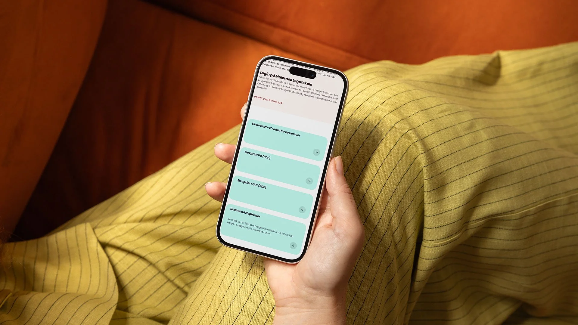 Person, der holder en smartphone med en skolestart-website åben, sidder på en gulstribet bukser, og sofa i baggrunden.